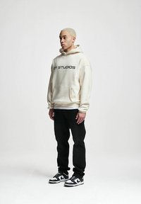 Felpa crema con testo "2Y STUDIOS", pantaloni sportivi neri e sneakers bianco e nere. La felpa presenta una tasca frontale e una vestibilità comoda.