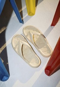 Tongs beige reposant sur un sol blanc parmi les pieds de chaises en plastique rouges, bleues et jaunes projetant des ombres.
