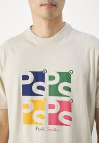 Beige Baumwoll-T-Shirt mit einem Grafikdesign von vier farbigen Quadraten—blau, grün, gelb und pink—die jeweils mit stilisierten Texten und Logo-Akzenten versehen sind.