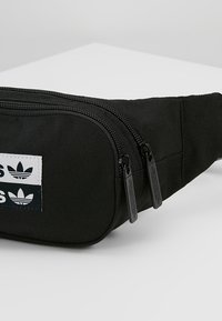 Černá plátěná ledvinka s dvojitými zipovými přihrádkami, bílou nášivkou s černým textem a designem tří listů, a nastavitelným popruhem.