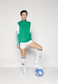 Grøn og hvid atletisk top med lynlås i kraven, parret med hvide shorts og knæhøje sokker med tre sorte striber. Hvide fodboldstøvler og en farverig fodbold.