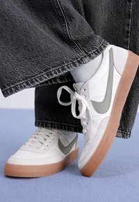 Tênis atléticos brancos com detalhes cinzas, parte superior em couro liso, sola texturizada e bico redondo. Combinados com calças de denim preto soltas.