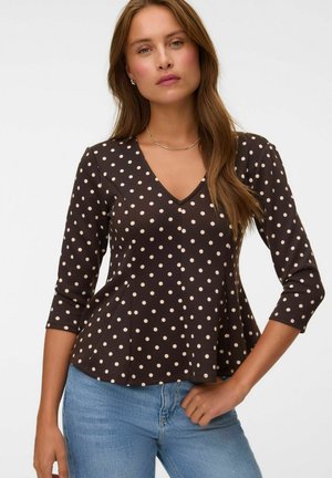 Femme portant un haut noir à manches longues à pois blancs et un jean bleu, debout devant un fond blanc uni.