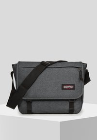Borsa a messaggero grigia con tessuto testurizzato, tracolla nera regolabile, chiusura a pattina e patch del logo rosso. Presenta una tasca con zip sul lato.