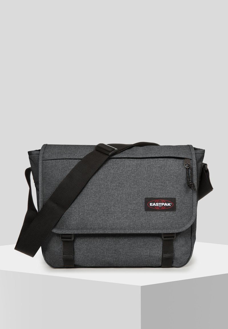 Borsa a messaggero grigia con tessuto testurizzato, tracolla nera regolabile, chiusura a pattina e patch del logo rosso. Presenta una tasca con zip sul lato.