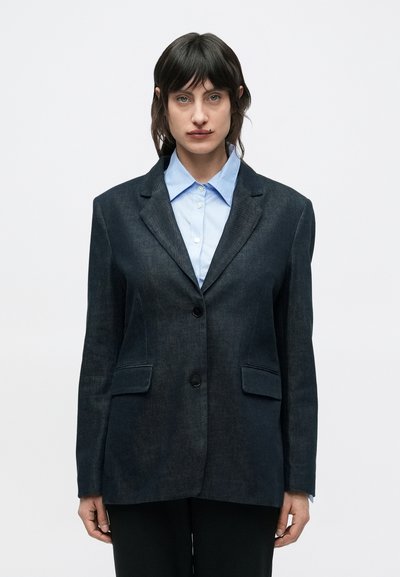 Blazer en denim foncé à coupe ajustée, fermeture à un bouton, revers crantés et deux poches avant. Porté sur une chemise bleu clair.