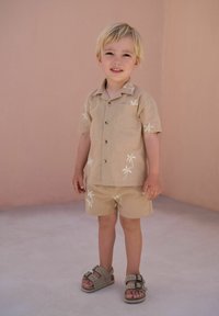 Maglietta a maniche corte color tan e pantaloncini abbinati, entrambi con ricami bianchi di palme. Il bambino indossa sandali grigi con due fibbie. Sfondo neutro.