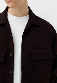 Ciemnobrązowa kurtka z guzikami, z dwiema kieszeniami na piersi i gładką teksturą, noszona na białym t-shircie z okrągłym dekoltem.