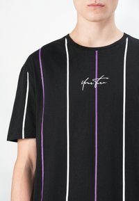 Camiseta negra con rayas verticales blancas y moradas, con un logo manuscrito "You Then" en el pecho, fabricada en un material de algodón suave.