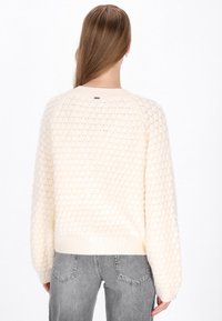 Sweter w kolorze kremowym z teksturowanym wzorem w plastry miodu, ściągaczem przy kołnierzu i u dołu oraz długimi rękawami. W komplecie z szarymi dżinsami.
