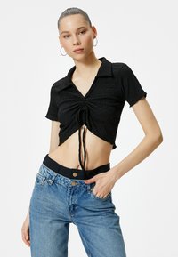 Sort cropped skjorte med krave, struktureret stof og knude foran. Båret med højttaljede lyseblå denimjeans.