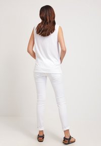 Vit ärmlös topp i bomull, kombinerad med åtsittande vita byxor och bruna sandaler, sedd bakifrån mot en neutral bakgrund.