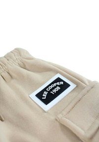 Shorts en tissu beige avec ceinture élastique, comportant une étiquette noire et blanche indiquant "LEE COOPER 1908". Texture lisse, design simple.