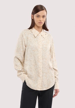 Kobieta w beżowej, wzorzystej koszuli z długim rękawem i szerokim kołnierzem oraz czarnych spodniach, stoi z rękami w kieszeniach na jednolitym tle.