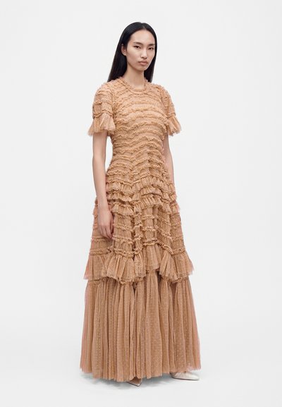 Femme aux longs cheveux noirs portant une robe beige texturée longue jusqu'au sol avec des volants et des manches courtes, debout devant un fond blanc uni.