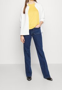 Vit skjorta med knappar över en gul t-shirt, i kombination med blå raka jeans och svarta skor med metalliska detaljer.