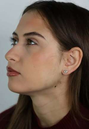 Jonge vrouw met lang bruin haar die omhoog kijkt, gekleed in een wijnkleurige top en een kleine diamantstud in haar linkeroor.