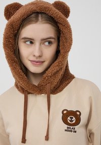 La felpa marrone con cappuccio presenta un cappuccio a forma di orsetto morbido, con orecchie, un corpo crema e un viso di orso ricamato con il testo "RELAX MOOD ON."