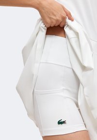 Main levant un vêtement blanc pour dévoiler un short blanc ajusté avec un petit logo crocodile vert brodé sur la cuisse.