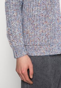 Sweter kardigan w jasno niebieskim kolorze z wielobarwnymi drobnymi plamkami. Posiada żebrowane mankiety i dół oraz kieszeń z przodu. Miękki, o fakturze materiał.