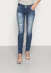 Jean skinny bleu avec des zones déchirées et décolorées, en denim ; comporte cinq poches et un zip à bouton. Porté avec des sandales à talons beiges.