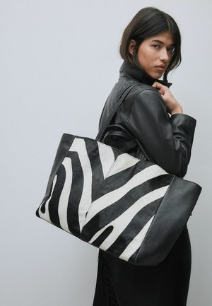 Žena v čiernej koženej bunde drží veľkú čierno-bielu tašku s pásikmi zebry cez rameno na jednofarebnom pozadí.