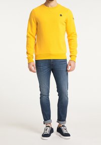 Felpa gialla con polsini e orlo a coste, con logo nero sul petto, abbinata a jeans aderenti blu e sneakers blu navy.