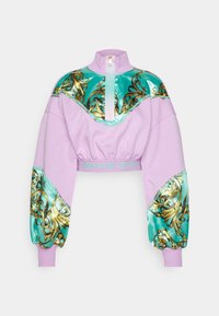 Avskuren sweatshirt i lavendel med hög krage, med turkosa blanka detaljer och gyllene barockmönster på ärmarna. Gyllene dragkedjedetalj.