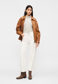 Kobieta w brązowej skórzanej kurtce, w paskowanym golfie, białych spodniach szerokich u dołu i brązowych botkach, stojąca na jednolitym tle.