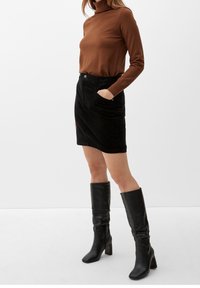 Brauner Rollkragenpullover, schwarzer Cordminirock mit Taschen und knielangen schwarzen Lederstiefeln mit Absatz. Glatte Texturen und gerader Schnitt.