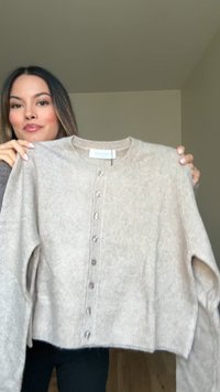Frau, die einen beigen, langärmligen Cardigan mit Knöpfen in einer schlichten Innenumgebung hält.
