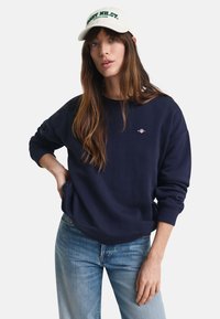 Marine blå hettegenser med ribbestrikkede mansjetter og tilpasset linning, med en liten logo på brystet. Sammen med lyseblå jeans og en kremfarget baseballcap.