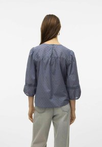 Modrá blůza s nabíranými rukávy, texturovaným vzorem, kulatým výstřihem a krajkovými detaily; doplněná o světle šedé džíny volného střihu.