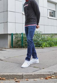 Person som bär en mörk sweatshirt, blå jeans och vita sneakers, står på en trottoar nära en byggnad med ett grönt staket och växter.