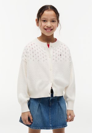 Ung pige smilende, iført en hvid knapstrikket cardigan med mønstre over en rød skjorte og et blåt denimskørt.