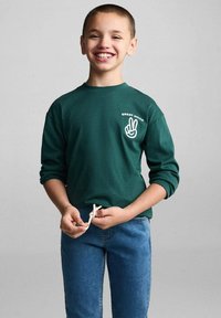 Tmavě zelené tričko s dlouhým rukávem a nápisem "SKVĚLÉ NÁLADY" spolu s grafikou mírového symbolu; v kombinaci s modrými džíny a detailem na šňůrku.