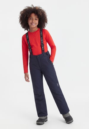 Laps lokkis juustega, seljas punane pika varrukaga särk, tumesinised lumepüksid trukkidega ja mustad talvesaapad, naeratab, üks käsi taskus.