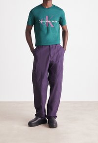 Zelené bavlněné tričko s grafikou "Calvin Klein Jeans", spárované s fialovými cargo kalhotami a černými nazouvacími botami. Neformální outfit s kapsami.