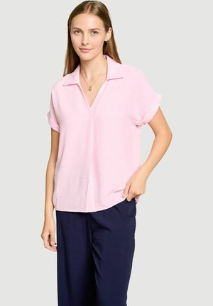 Jeune femme aux cheveux lisses châtain clair portant un chemisier rose clair à manches courtes et un pantalon bleu marine foncé sur un fond uni.