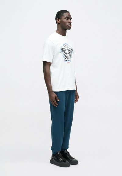 Valge T-särk, mille ees on siniste detailidega kaunistatud näo graafika, koos lahtiste tumeroheliste pükstega ja mustade vabaajajalatsitega.