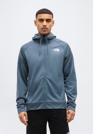 Mann trägt eine blaue Kapuzenjacke mit Reißverschluss, Kapuze und Logo auf der Brust, steht mit entspannten Händen an den Seiten vor einem einfarbigen Hintergrund.