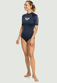 Námořnická modrá rash guard s krátkým rukávem a přiléhavým střihem, s bílým logem na hrudi a rukávu, doplněná o odpovídající bikiny.