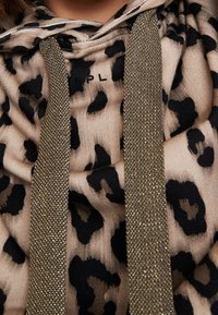 Felpa con cappuccio beige con stampa leopardata nera. Presenta lacci dorati lucidi e testurizzati e un tessuto morbido. Logo del marchio visibile.