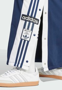 Marina och vita Adidas joggingsbyxor med tre vita ränder, logotypklistermärken och knappstängning; matchad med vita Samba-sneakers med gummisulor.
