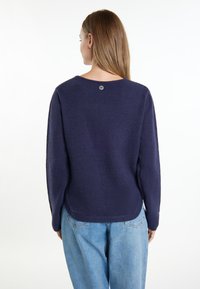 Marineblauer Pullover mit weicher Textur, rundem Halsausschnitt und einem Knopfdetail am Rücken. Kombiniert mit locker sitzenden hellblauen Jeans.