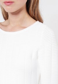 Pull blanc texturé avec un motif côtelé, doté d'un large col et d'un tissu doux. Gros plan sur l'épaule et la zone du col.