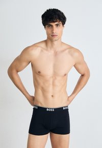 Schwarze Baumwoll-Boxershorts mit einem weißen "BOSS"-Logo am Bund; das Model steht mit Händen in die Hüften gestützt und hat einen neutralen Gesichtsausdruck.