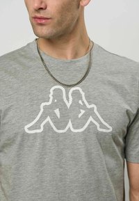 Nærbilde av en person som har på seg en grå T-skjorte med en hvit logo av to rygg-til-rygg sittende silhuetter og et sølvkjede.