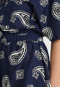 Tissu bleu marine avec motifs cachemire blancs et noirs et motifs géométriques, avec taille élastique froncée et manche courte sur le bras visible.