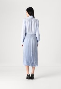 Vestido plissado azul claro com mangas longas, gola alta e cintura franzida. Usado com sapatos pretos de salto. Textura de tecido suave.